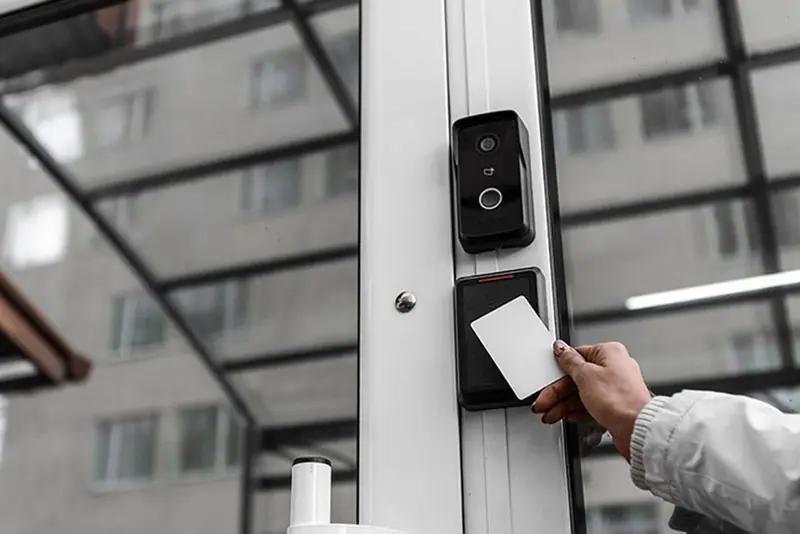 Hand holding a key card to a reader below a video intercom on a glass building entrance