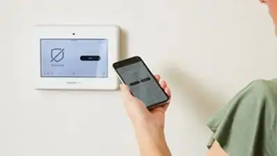 Person holding a smartphone app in front of a wall-mounted touchscreen alarm panel